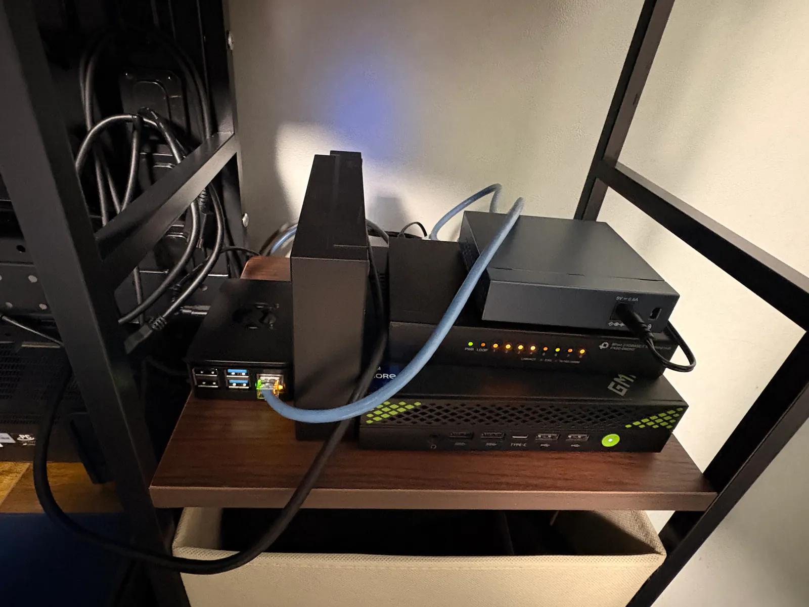 棚に積んだホームラボの機器。ミニPC、LEDが光るネットワークスイッチ、ケースに入った小型ボード、外付けドライブ