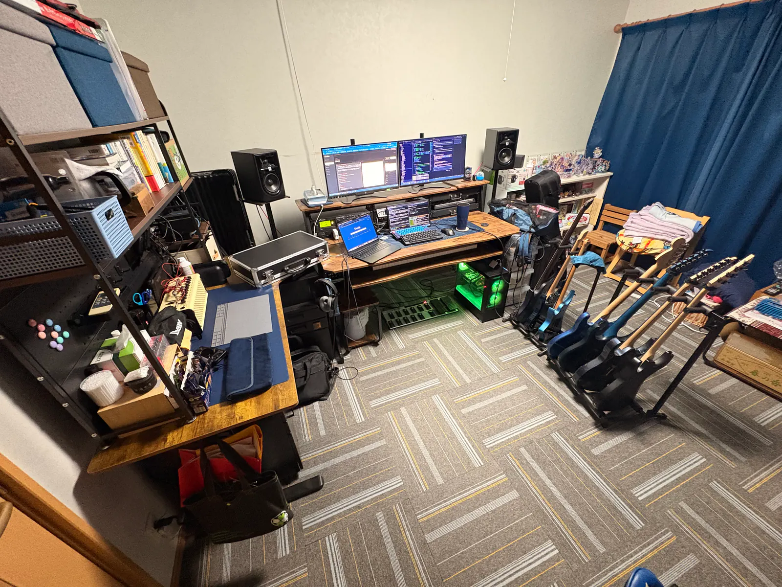作業部屋の全景。奥にモニター2台のデスク、右にスタンドに並ぶエレキギター、左に棚付きのサブデスク